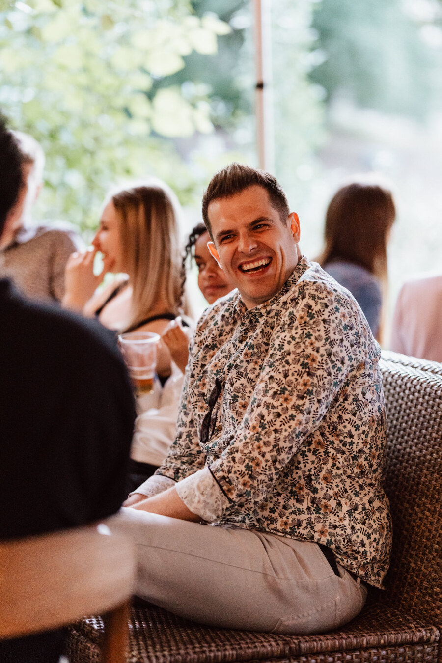 A man in a floral shirt sits on a wicker chair, smiling and laughing with others at an outdoor gathering. The people around him seem relaxed and engaged in conversation. 