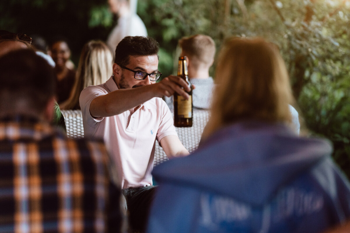 A man in a bright pink polo shirt and glasses picks up a bottle of beer while sitting outside with a group of people, greenery visible in the background.