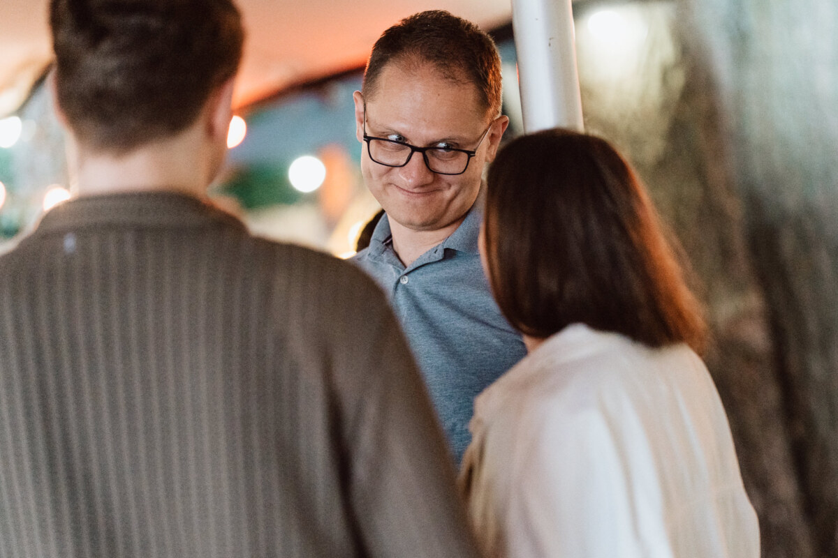 A man wearing glasses and a blue shirt smiles while talking to two people, one in a striped shirt and the other in a white top, at an outdoor meeting with colorful lights in the background.