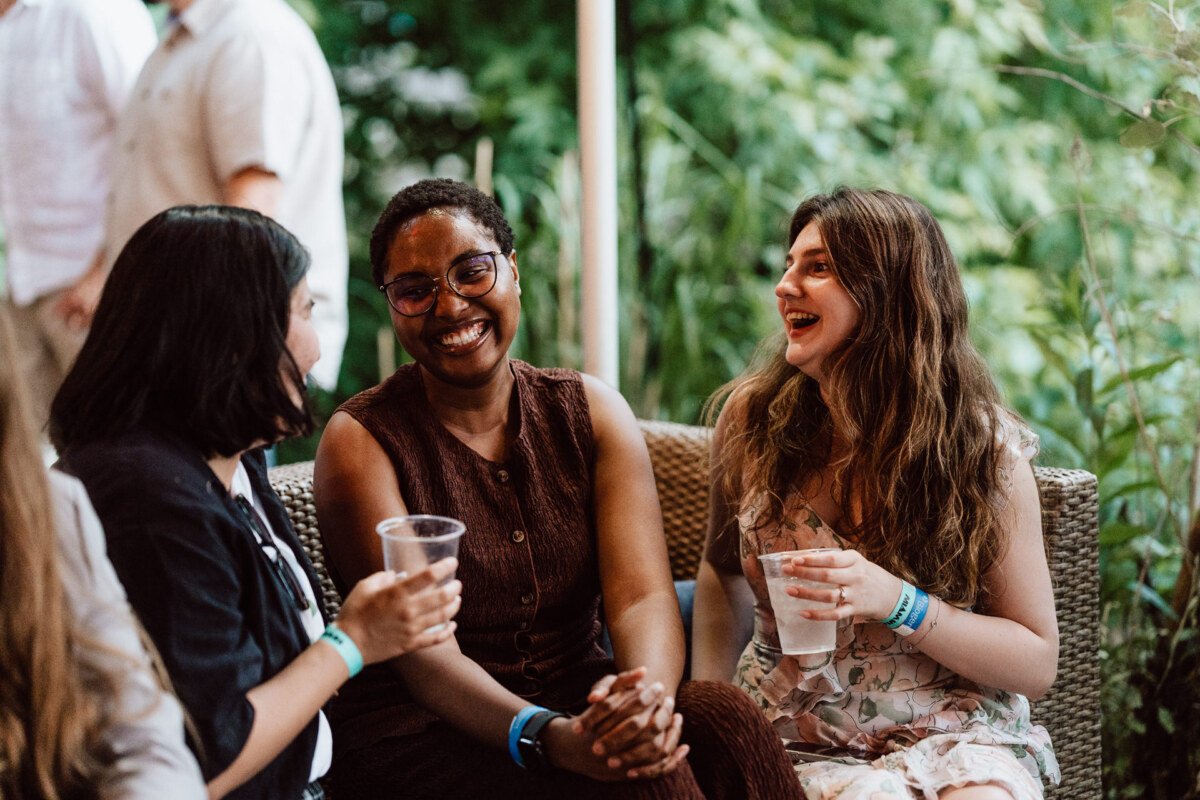 Three women sit close to each other on a wicker bench, holding drinks and laughing, enjoying relaxed conversation outdoors with the greenery in the background.