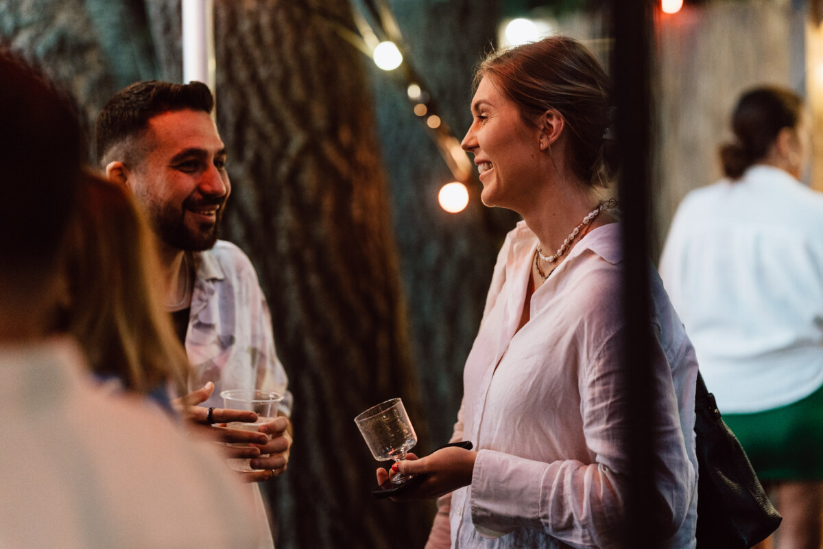 A man and a woman smile and talk while holding drinks at an outdoor meeting, with string lights and a tree trunk in the background.