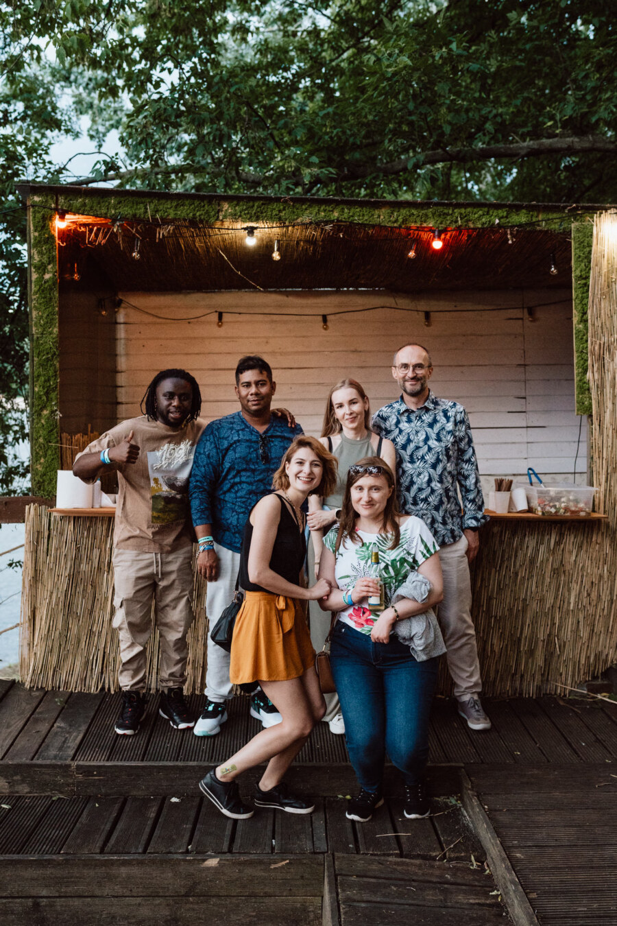 Six people smile and pose together in front of a rustic outdoor bar with a thatched roof, surrounded by greenery. Two women stand in front, four people stand behind them. The atmosphere seems friendly and relaxed.  