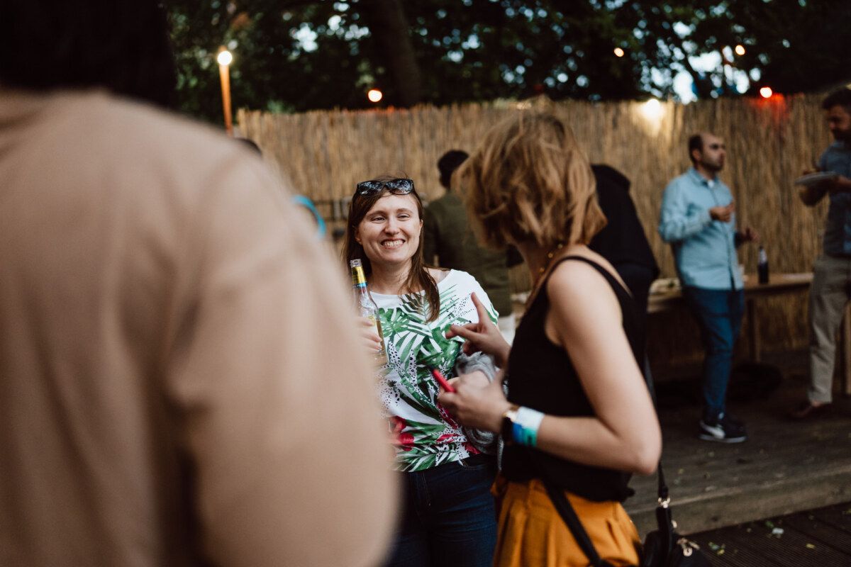 A woman smiling and holding a drink chats with another woman at an outdoor meeting with social people in the background. String lights and trees create a relaxing atmosphere. 