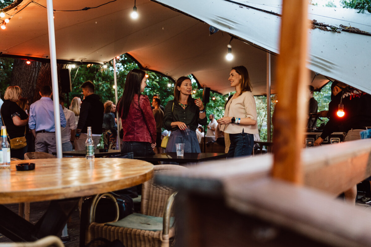 People gather under a large tent with string lights at an outdoor event. Three women stand in the foreground, talking, while others gather in groups behind them. Wooden tables and chairs are scattered around.  