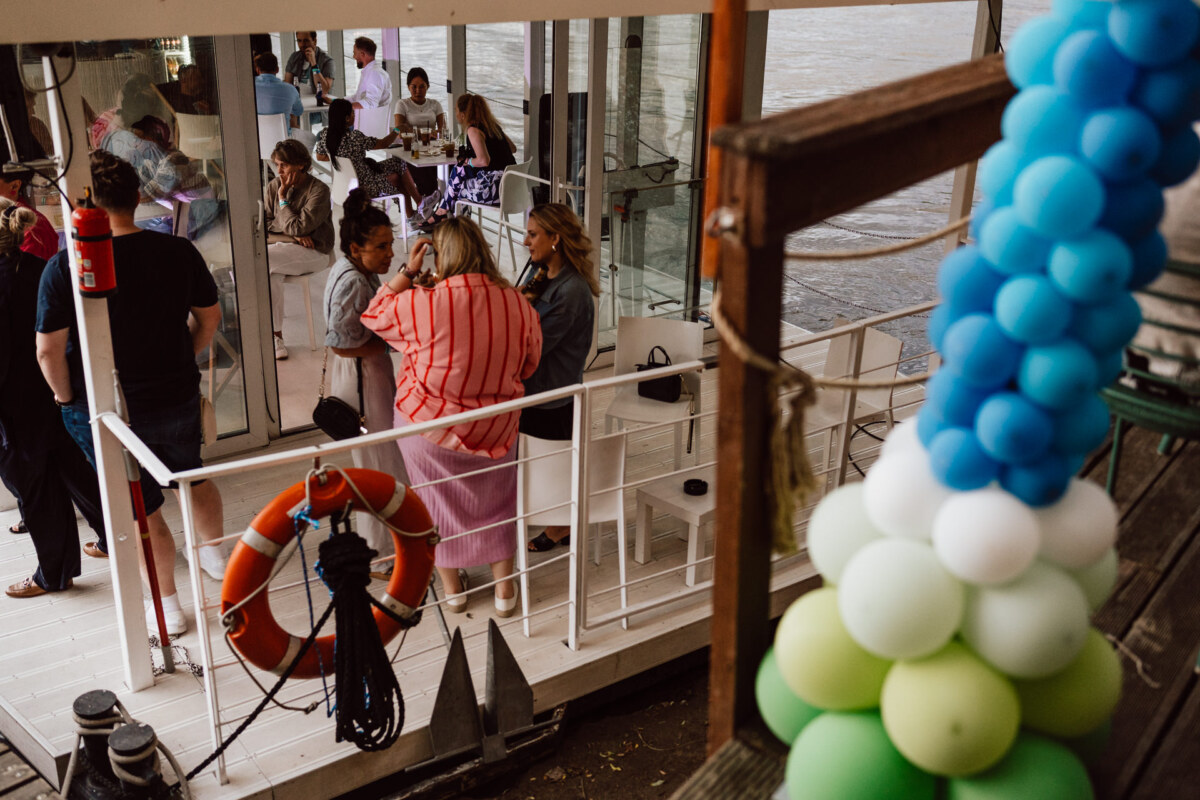 A group of people meet on the deck of a boat by the water. Blue and green balloons decorate the foreground, and a lifebelt hangs from the railing. Some guests are sitting, while others are standing and talking.  