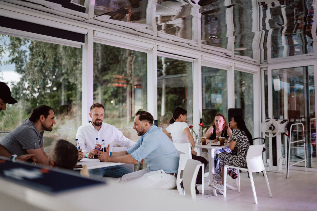 People are sitting and talking at tables in a bright, modern restaurant or cafe with glass walls. The scene looks relaxed with drinks on the tables, and trees are visible outside the windows. 