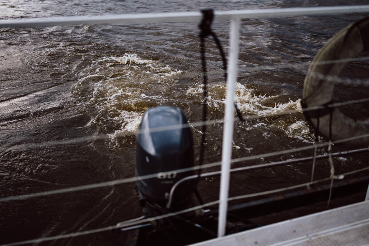 The boat's engine agitates the frothy water, viewed from behind a rope and net railing, creating swirling patterns on the river's surface.