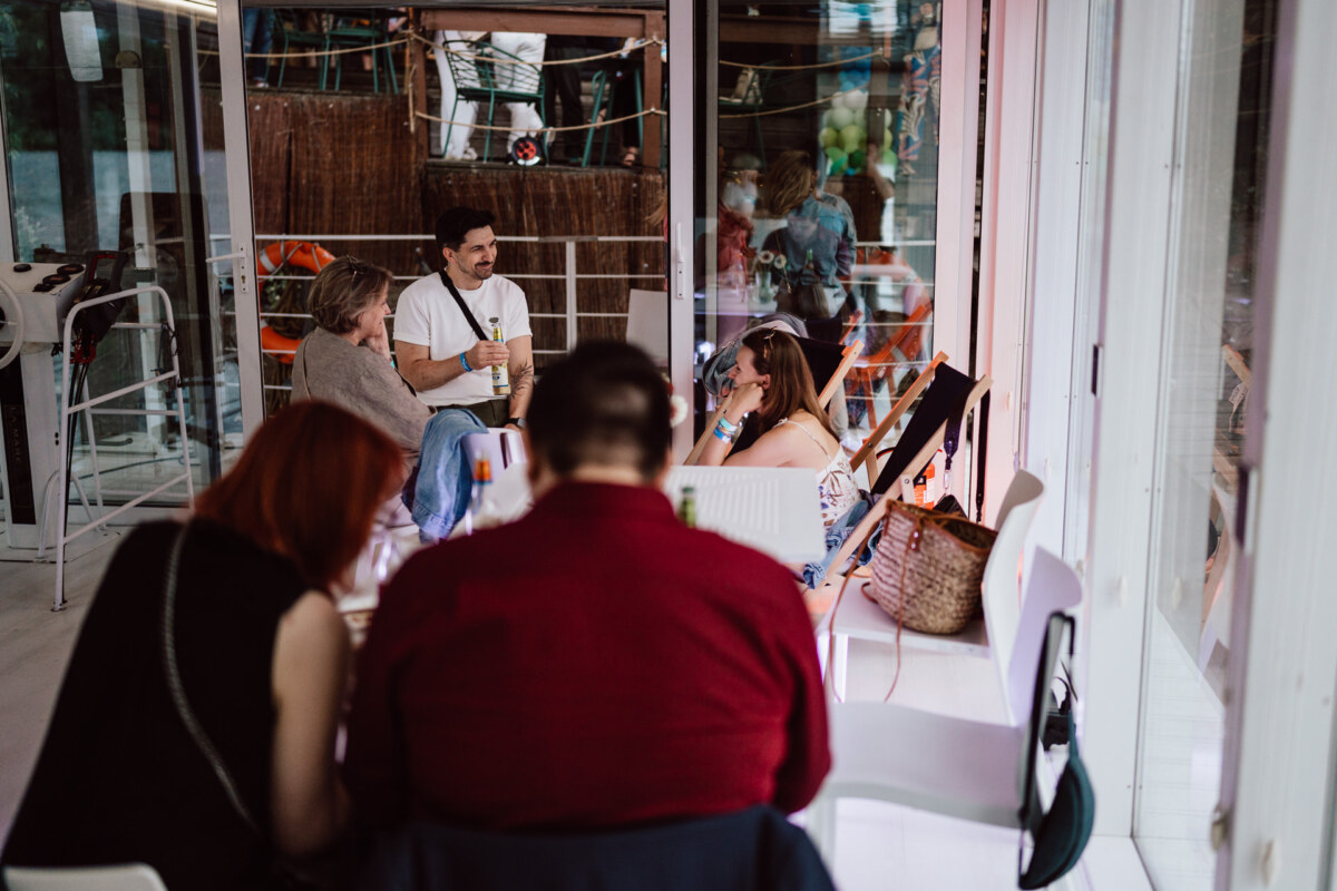 A group of people are sitting in a modern, glass-walled cafe. Some are talking and laughing, while others are focused on a laptop. The scene is casual and social, with natural light streaming in.  