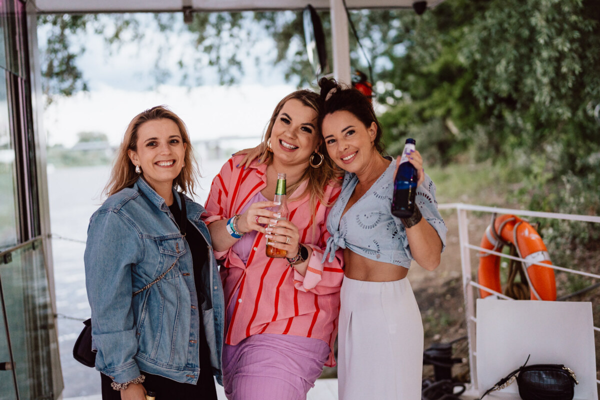 Three women are smiling and posing together outside, two holding bottles. One is wearing a denim jacket, another a pink and red striped shirt, and the third a light blue top and white skirt. Trees and water can be seen in the background.  
