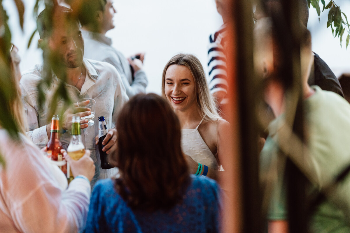 A group of people are meeting outdoors, holding drinks. A woman in a white top stands in the center, smiling radiantly, while others are gathered around her, engaging in conversation. Greenery and fuzzy elements surround the scene.  