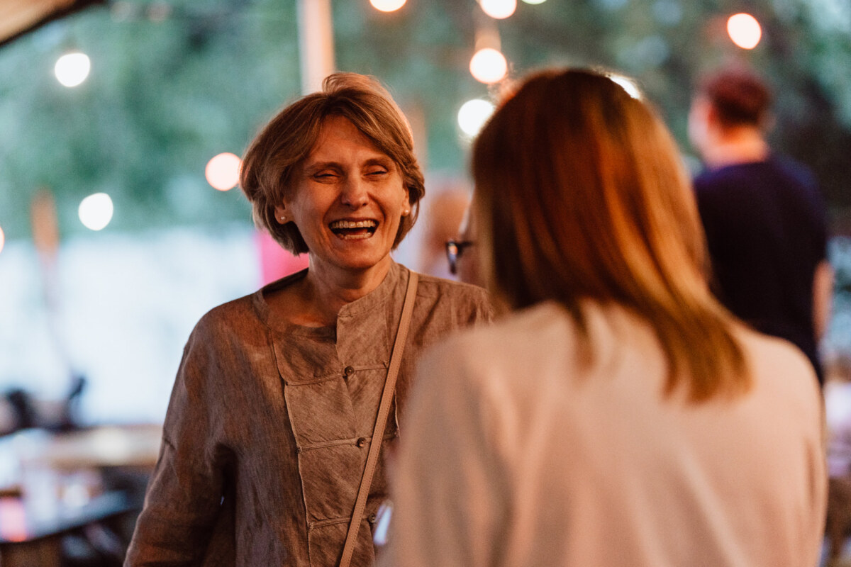 A woman with short hair smiles and laughs while talking to another person at an outdoor event with string lights and a blurry background.