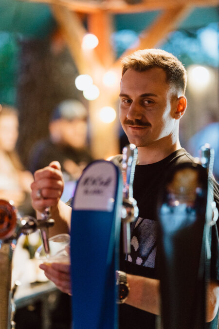 A mustachioed bartender smiles as he pours a drink from a tap at an outdoor bar, with colorful lights and fuzzy people in the background.