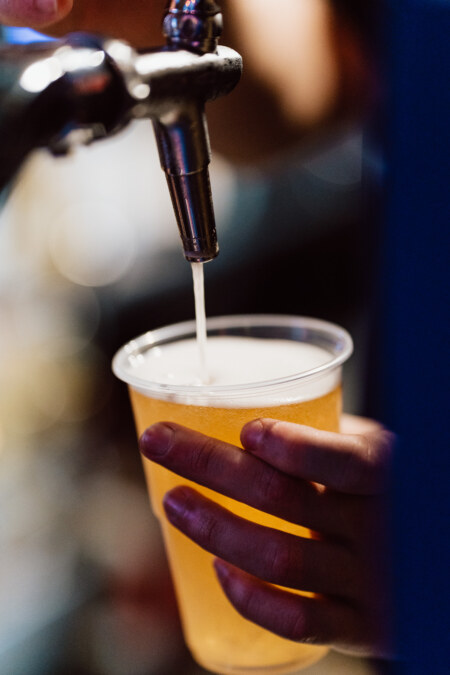 Close-up of a hand holding a plastic cup into which beer is poured from a metal tap, with foam forming on top of the golden liquid.