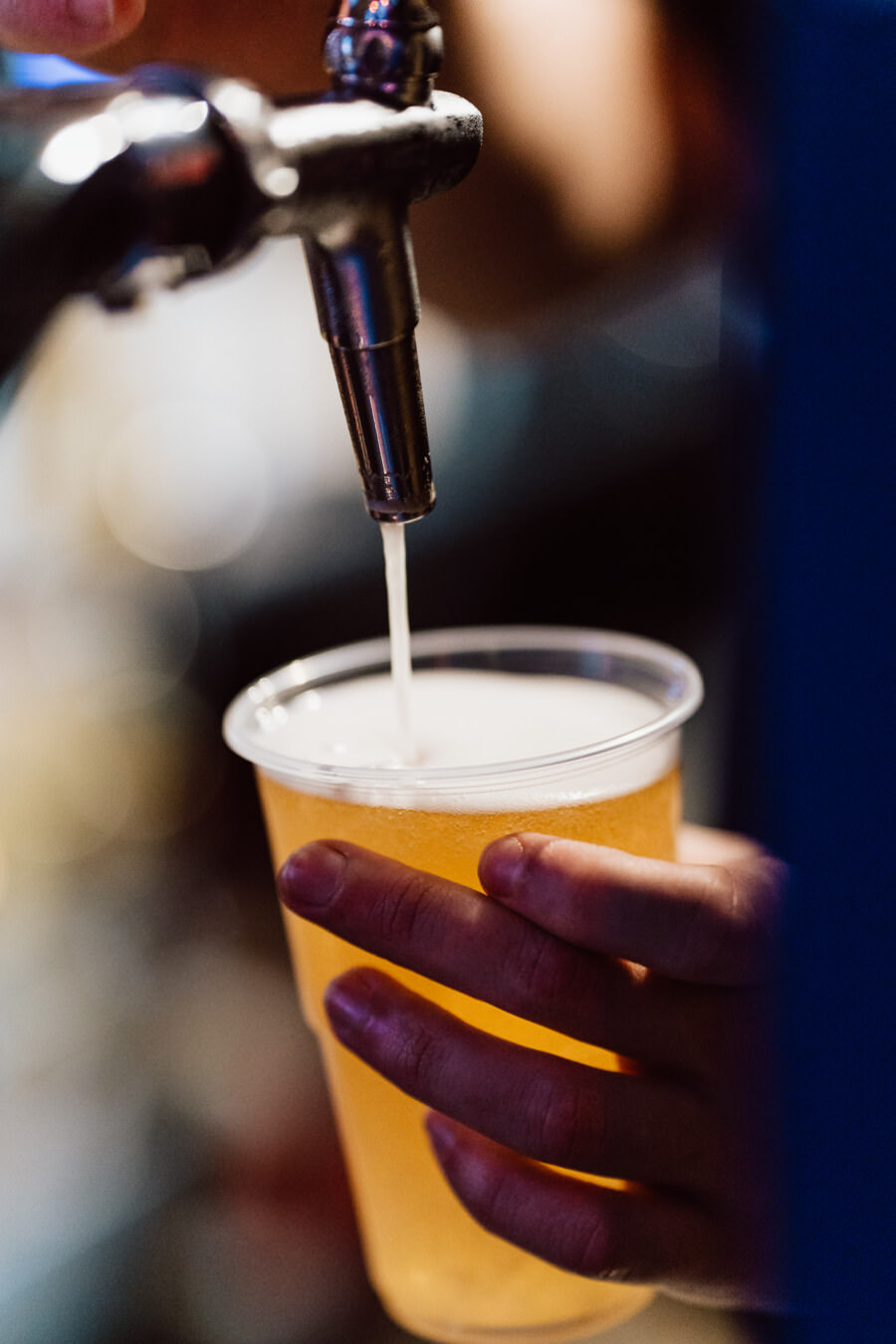 Close-up of a hand holding a plastic cup into which beer is poured from a metal tap, with foam forming on top of the golden liquid.