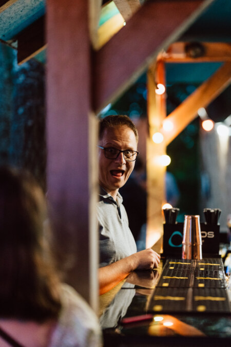 A man wearing glasses and with a puzzled expression is standing behind a bar counter outside in the evening, with colorful string lights and wooden beams in the background. An indistinct person can be seen in the foreground. 