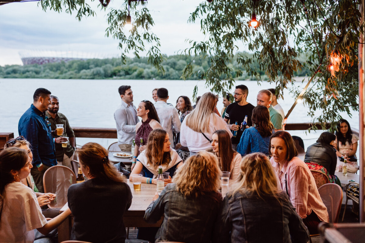 A group of people are meeting and having drinks on a patio by the river, surrounded by trees, with the stadium visible in the background. Some stand and talk, while others sit at wooden tables. 