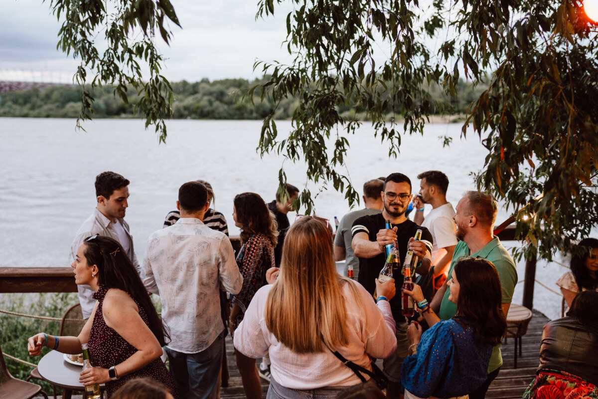 A group of people enjoy drinks and conversation at an outdoor meeting by the river, surrounded by trees, with a picturesque view of the water and greenery in the background.