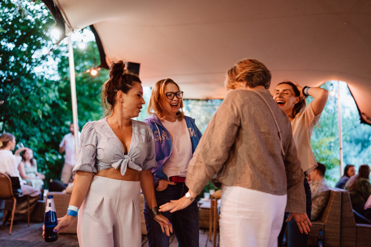 Four women laugh and dance together at an outdoor gathering under a tent, with trees and other people in the background. One of the women is holding a drink, and everyone seems to be having a good time. 
