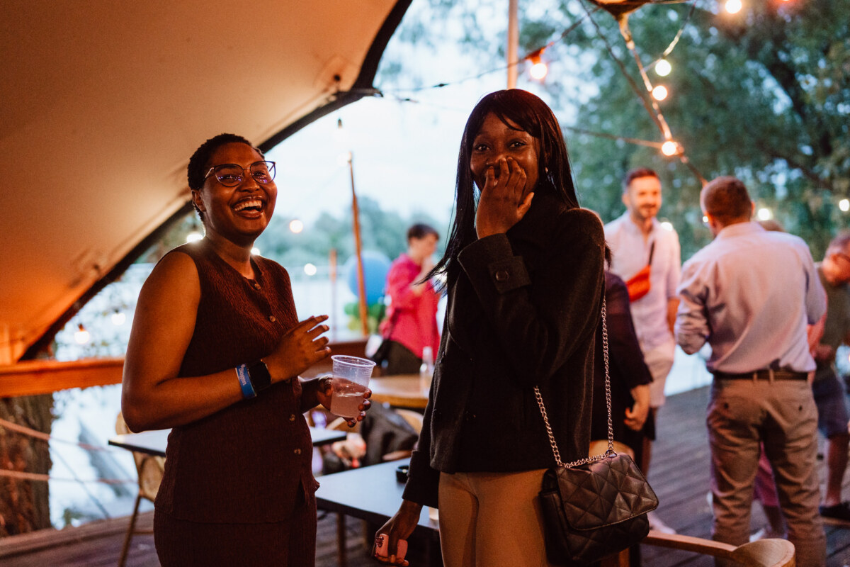 Two women smile and laugh together at an outdoor party under string lights while people mingle at tables in the background. One of the women holds a drink while the other puts her hand to her face, looking amused. 