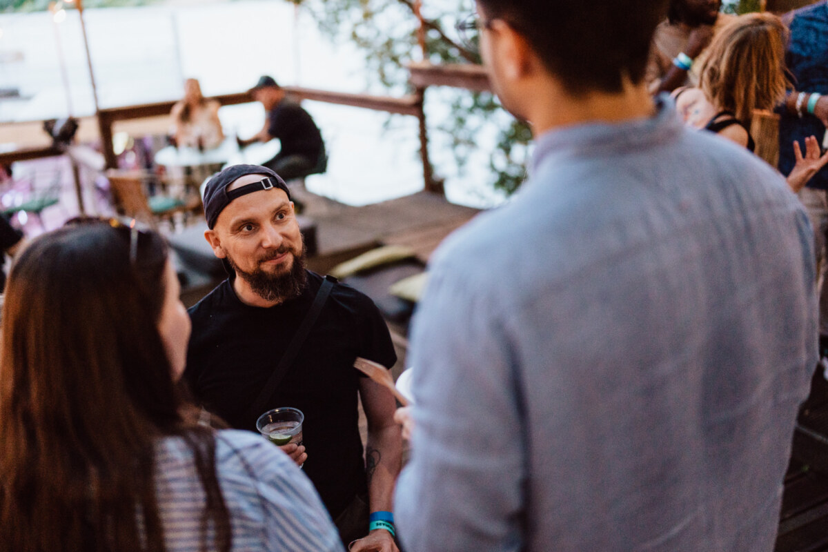 A bearded man wearing an upturned cap holds a drink while talking to two people at an outdoor social gathering near the water, with several other people in the background.
