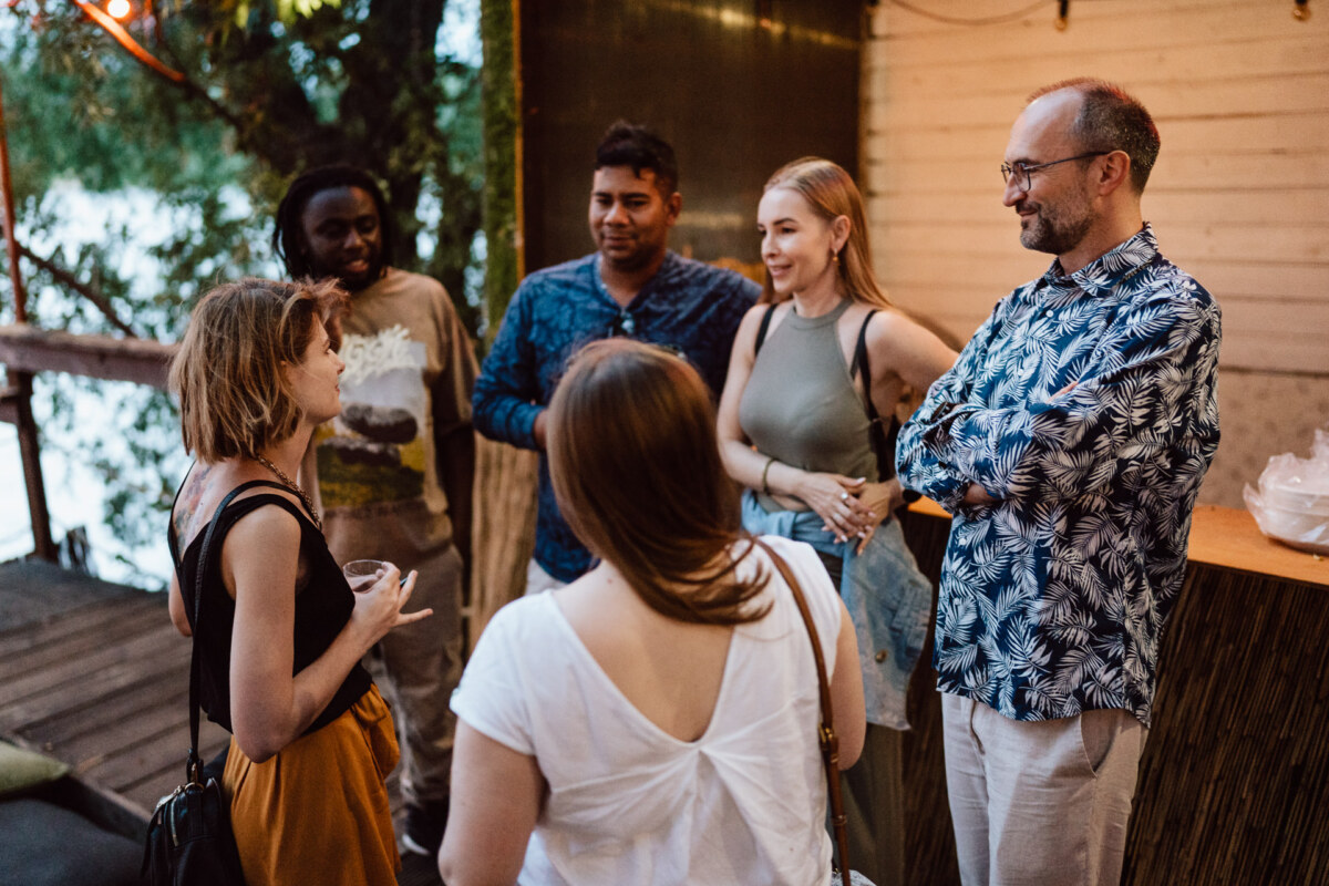 A group of six adults are standing outdoors having a conversation. Some are smiling and engaged, with trees and wooden walls in the background. One person is holding a drink.  