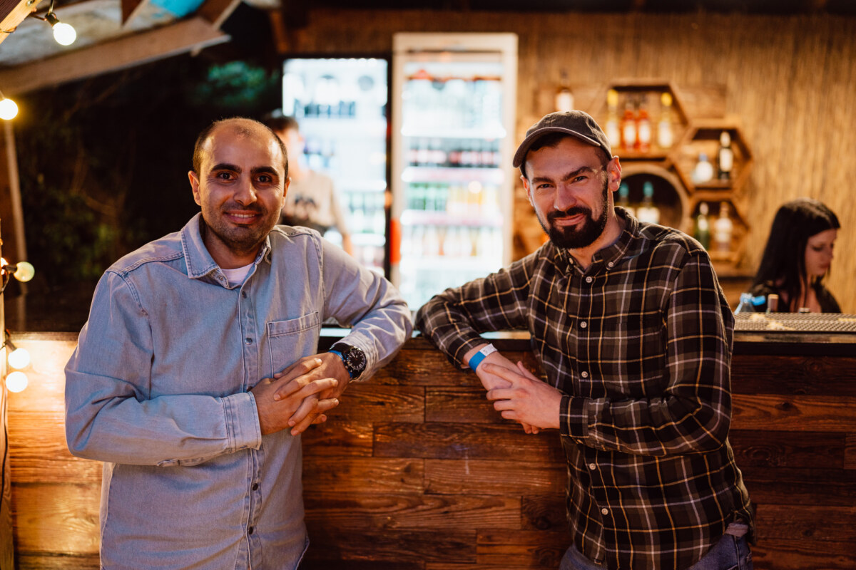 Two men are standing and smiling at each other at an outdoor wooden bar counter, with shelves of bottles and a refrigerator in the background. One of them is wearing a denim shirt, while the other is wearing a plaid shirt and a cap. A woman works behind the bar.  