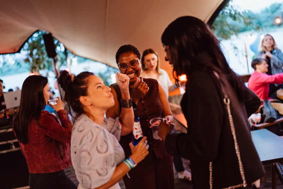 Three women stand together at a lively party, smiling and laughing, holding drinks. Other guests and warm lights are visible in the background, creating a casual and joyful atmosphere. 