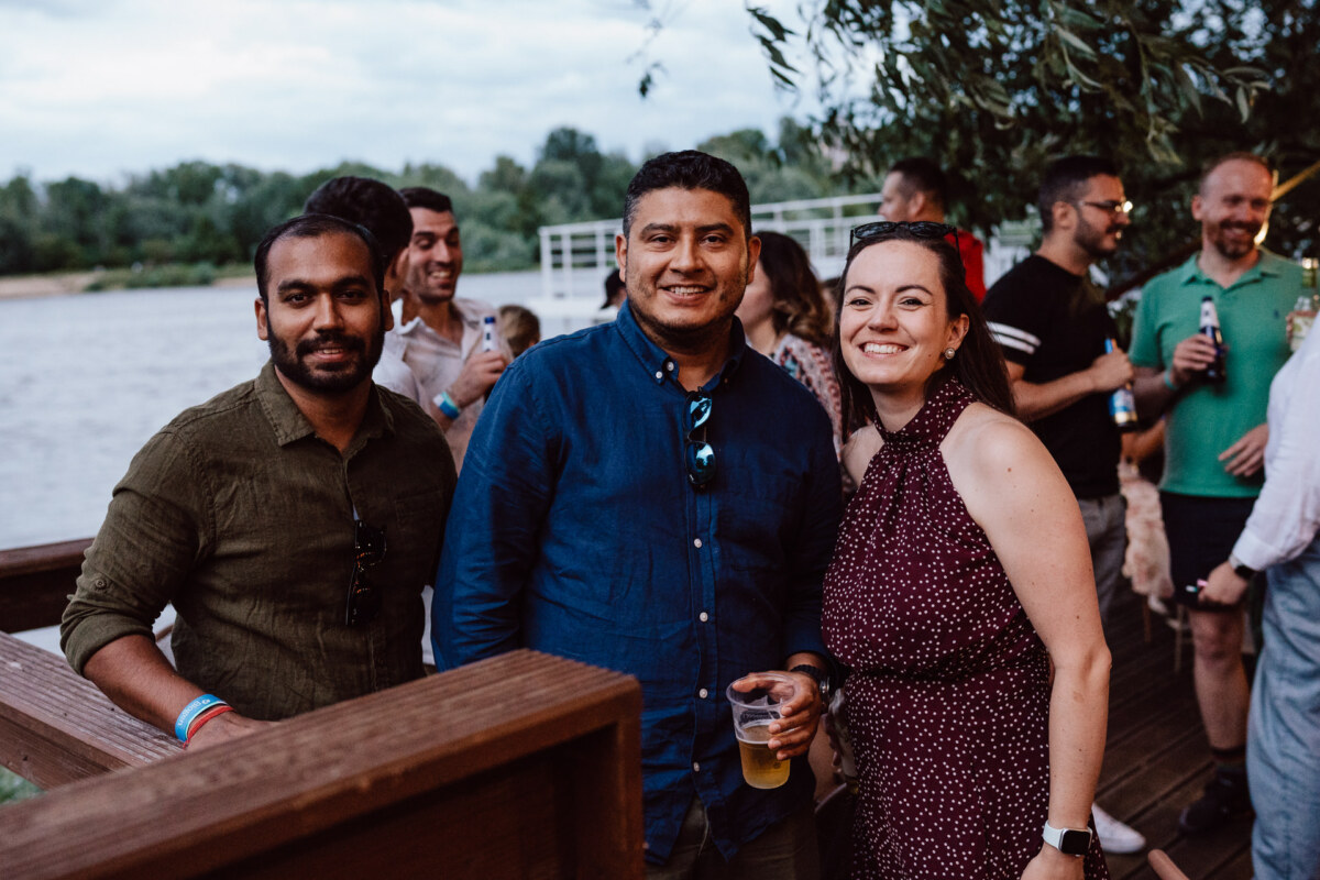 Three people smile for the camera on a wooden terrace by the water during an outdoor gathering. Other people in casual clothes talk and hold drinks in the background. Trees and water are visible.  