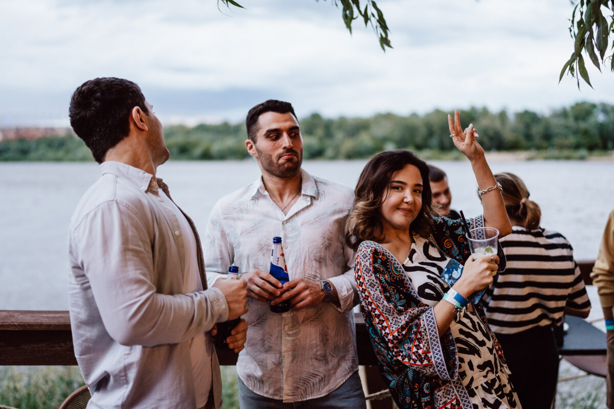 Three people are standing on the riverbank, two men are holding bottles, and a woman is smiling, raising her hand with a drink. Trees and water can be seen in the background, with more people partially visible behind them. 
