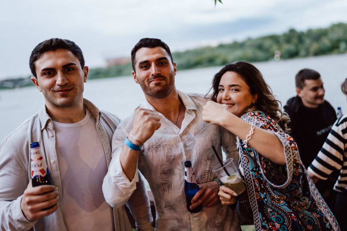 Three young adults are smiling and posing for a photo outdoors near the water. Two men hold bottles of beer and a woman with a drink playfully raises her fist, all looking cheerful during the casual encounter. 