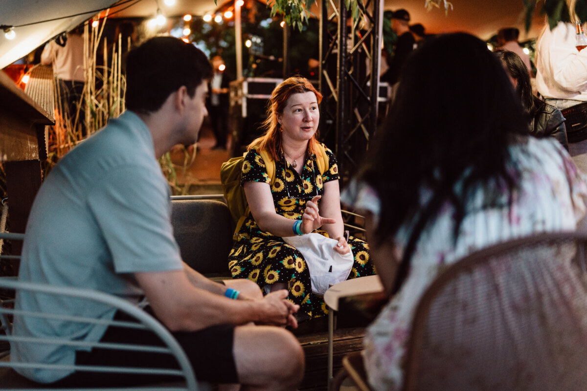 A woman in a dress with sunflowers sits and talks with two people during an outdoor evening gathering, surrounded by plants and string lights.