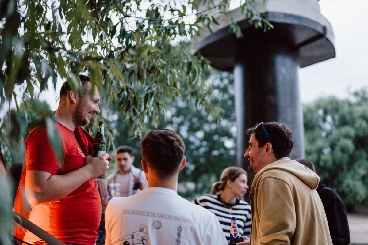 A group of people gather outdoors, talking and drinking drinks. Trees and a tall concrete structure are visible in the background. One person in a red shirt is talking to others, who are smiling and engaged in conversation.  