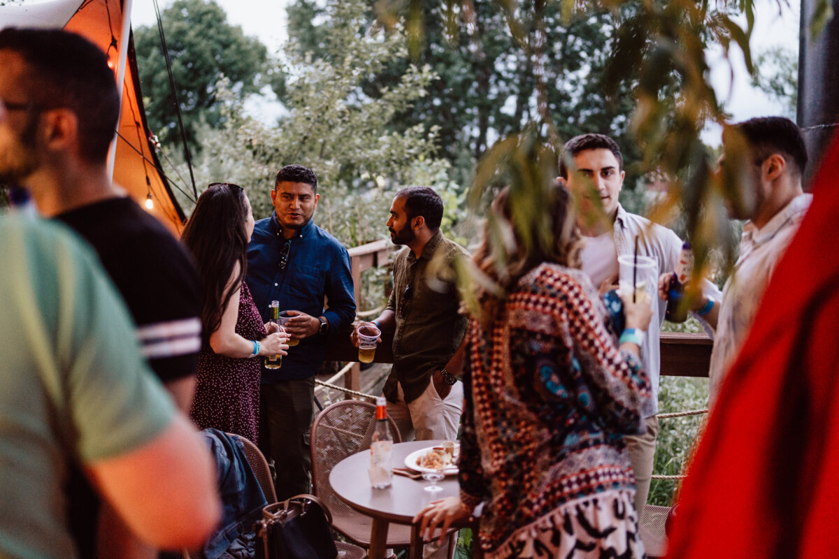 A group of people mingle and talk at an outdoor meeting, some holding drinks. There are trees in the background and a table with food and drinks in the center. The mood seems casual and social.  