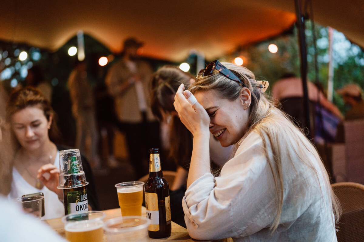 A woman sits at an outdoor table, laughing with her hand covering part of her face, surrounded by drinks and other people enjoying a social gathering in a warmly lit environment.