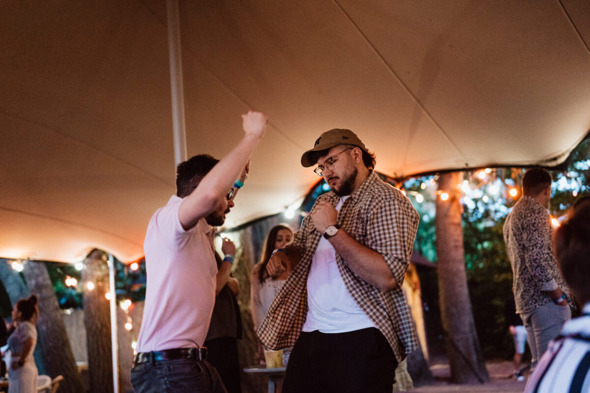 Two men dance enthusiastically under a canopy decorated with strings at an outdoor party. One of them is wearing a cap and plaid shirt, while the other is wearing a polo shirt. Other people and trees are visible in the softly lit background.  
