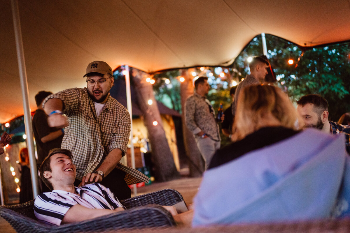 A group of people meeting outdoors under a canopy of string lights; one person standing playfully gestures toward a smiling friend sitting on a wicker chair, while others mingle in the background.