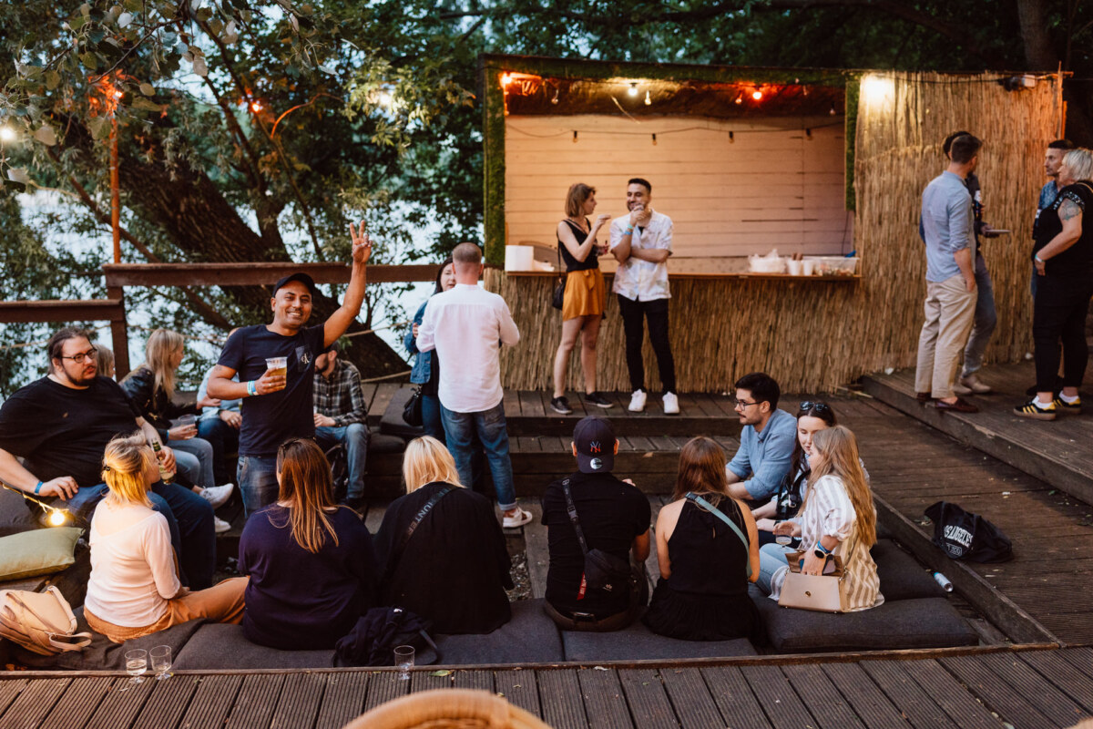 A group of people gather on an outdoor wooden patio, some sitting on cushions and others standing near the bar. Strings of lights and trees create a cozy, festive atmosphere. One person cheerfully waves to the camera.  