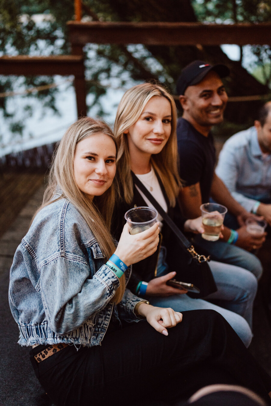 Three people are sitting close together outside, smiling at the camera and holding drinks in plastic cups, suggesting a casual social or party atmosphere. Trees and wooden railings are visible in the background. 