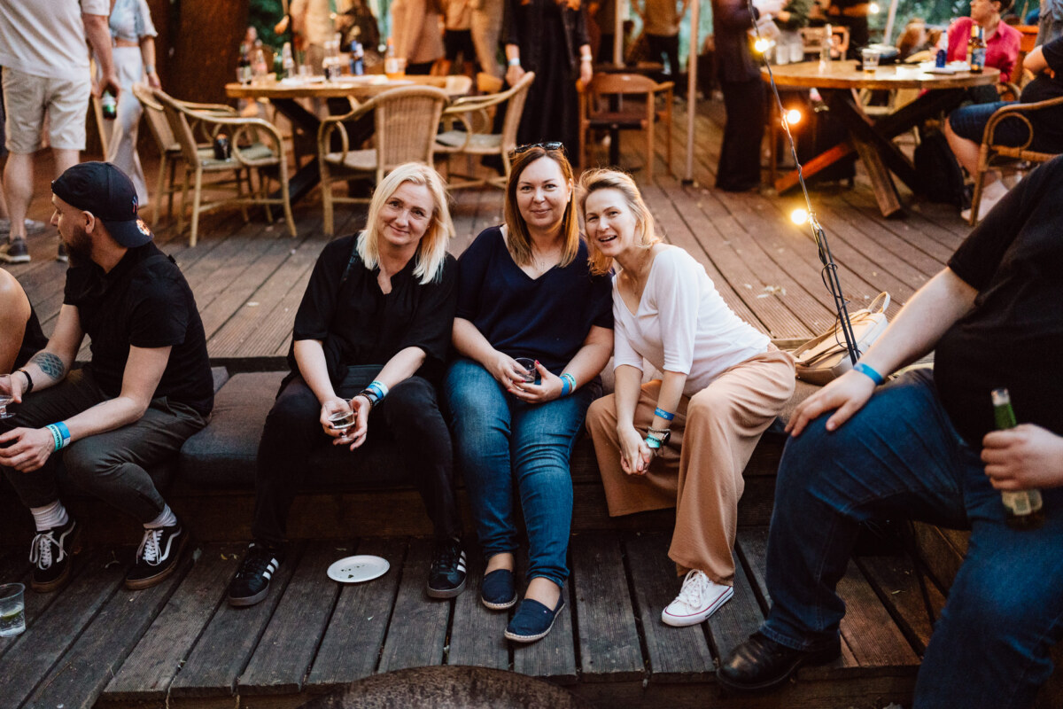 Three women sit close together on a wooden platform during an outdoor meeting, smiling at the camera. People are scattered, sitting and standing at wooden tables under string lights. 
