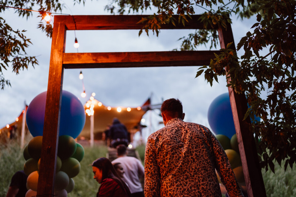 People walk under a wooden arch decorated with balloons toward a tent with string lights. Trees and greenery surround the area, suggesting an outdoor party at dusk. 