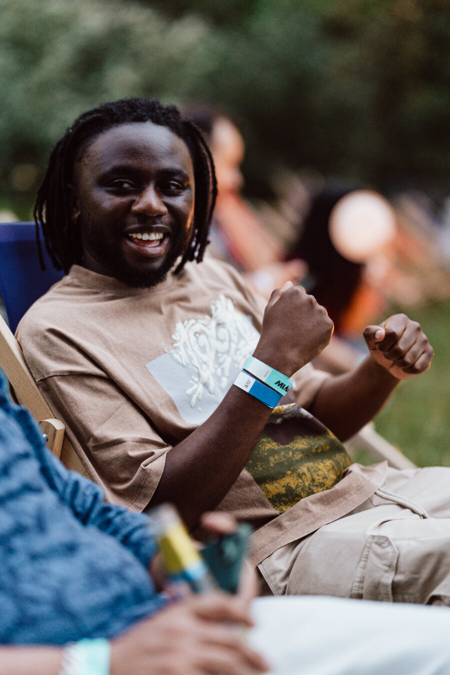 A smiling man with short dreadlocks sits outside in a chair, wearing a tan graphic T-shirt and wristbands, with a blurry background of greenery and people.