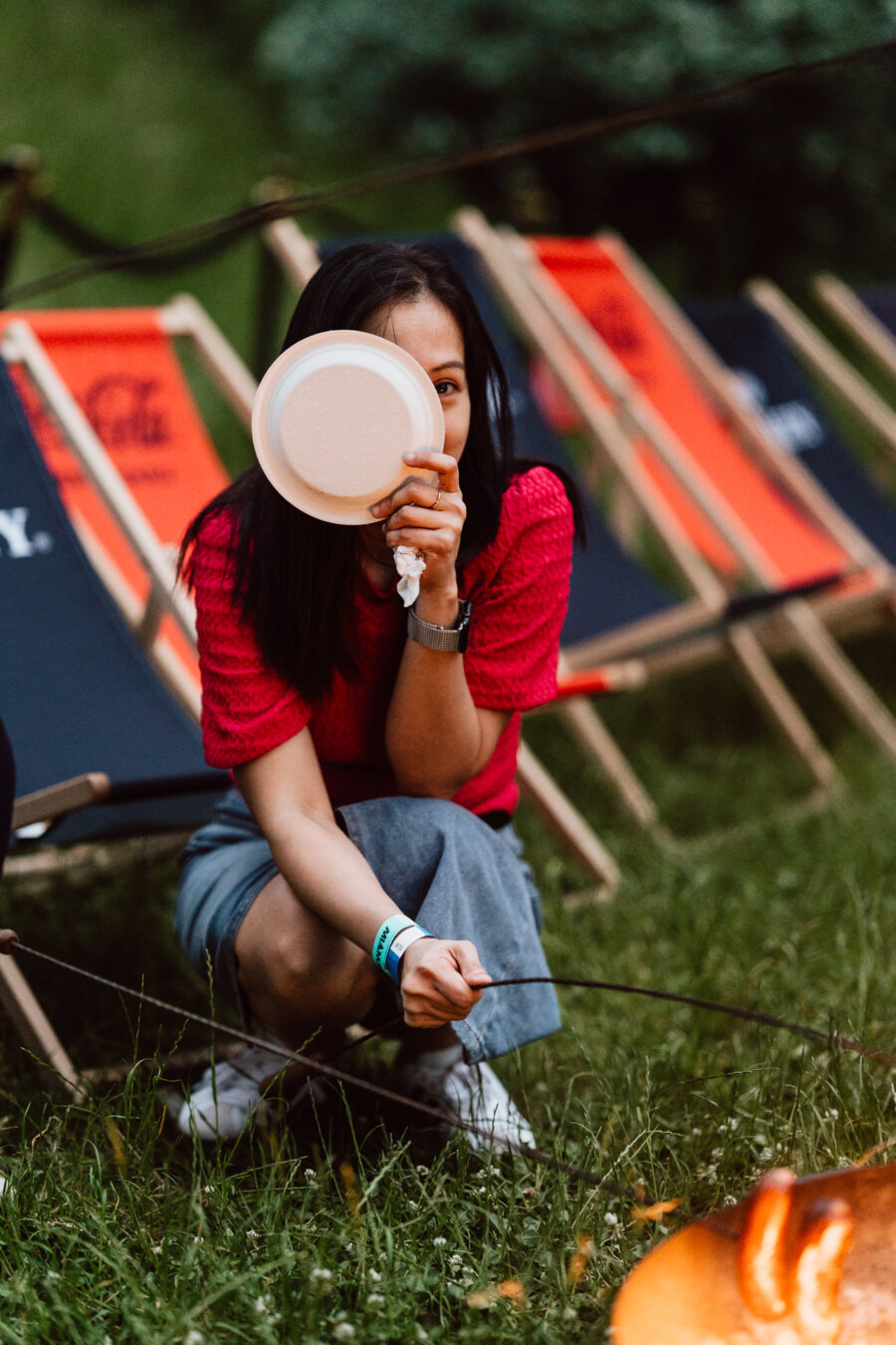 A woman in a red blouse and gray skirt sits on the grass in front of folding chairs, holding a white plate in front of her face and looking into the camera. She is outside, probably at a picnic or party. 