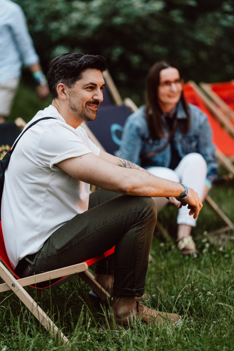 A man in a white shirt and olive pants sits on a red recliner outside, smiling. A woman in a denim jacket and white pants sits on a nearby chair. In the background is green grass and trees.  