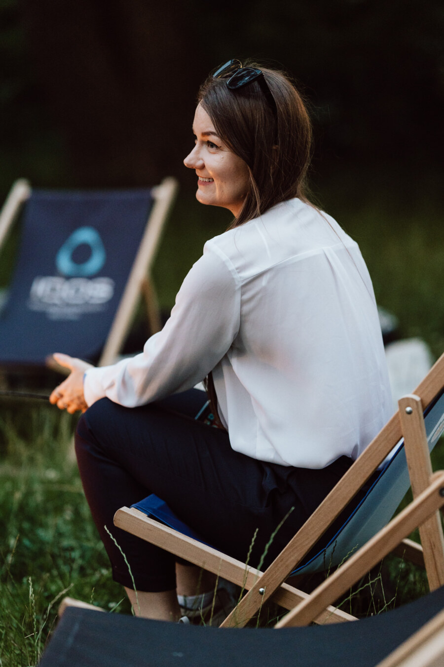 A woman with long brown hair, wearing a white blouse and dark pants, sits on a wooden deck chair outside. She smiles and looks to the side. The background is green with grass visible and another empty chair.  