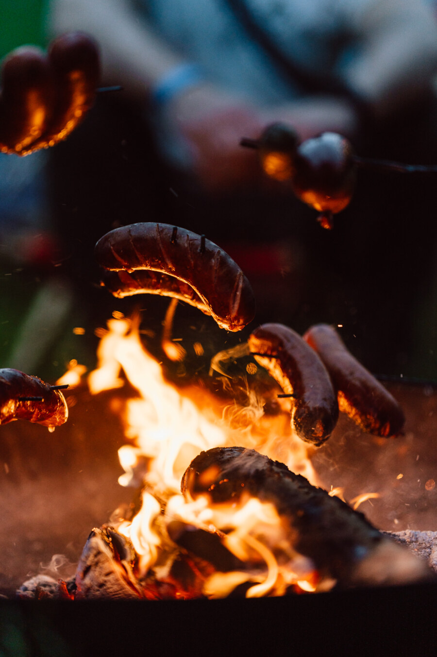 Sausages roasted on an open campfire, held on sticks over bright flames, with fuzzy figures in the background.