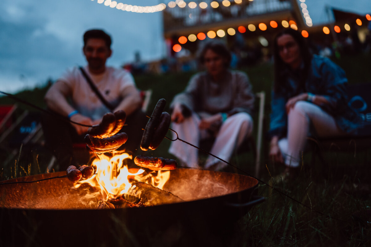 Three people sit around a campfire, roasting sausages on sticks. The scene takes place outside at dusk, with string lights and a building in the background, creating a cozy, festive atmosphere. 