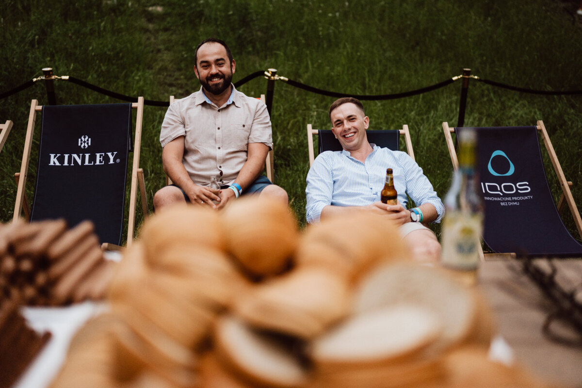 Two men sit and smile on deck chairs outside with the brand's logo behind them. In the blurred foreground are stacked wooden bowls and drinks on a table. Grass and a rope barrier are visible in the background.  