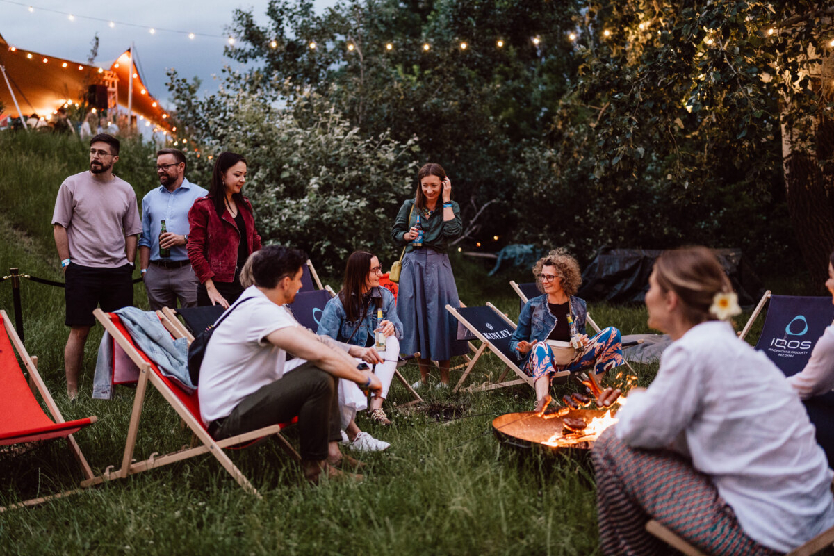 A group of people gather outside in the evening, sitting on deck chairs around a small campfire. Some are standing, talking and smiling. Strings of lights and greenery create a cozy, festive atmosphere.  