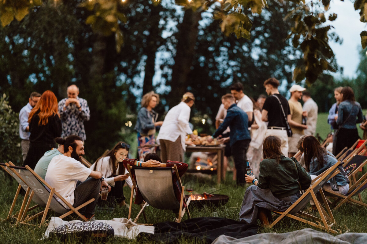 A group of people gather outdoors on a grassy area, sitting on folding chairs around a campfire, while others stand at tables with food, enjoying a relaxed evening social event under the trees.
