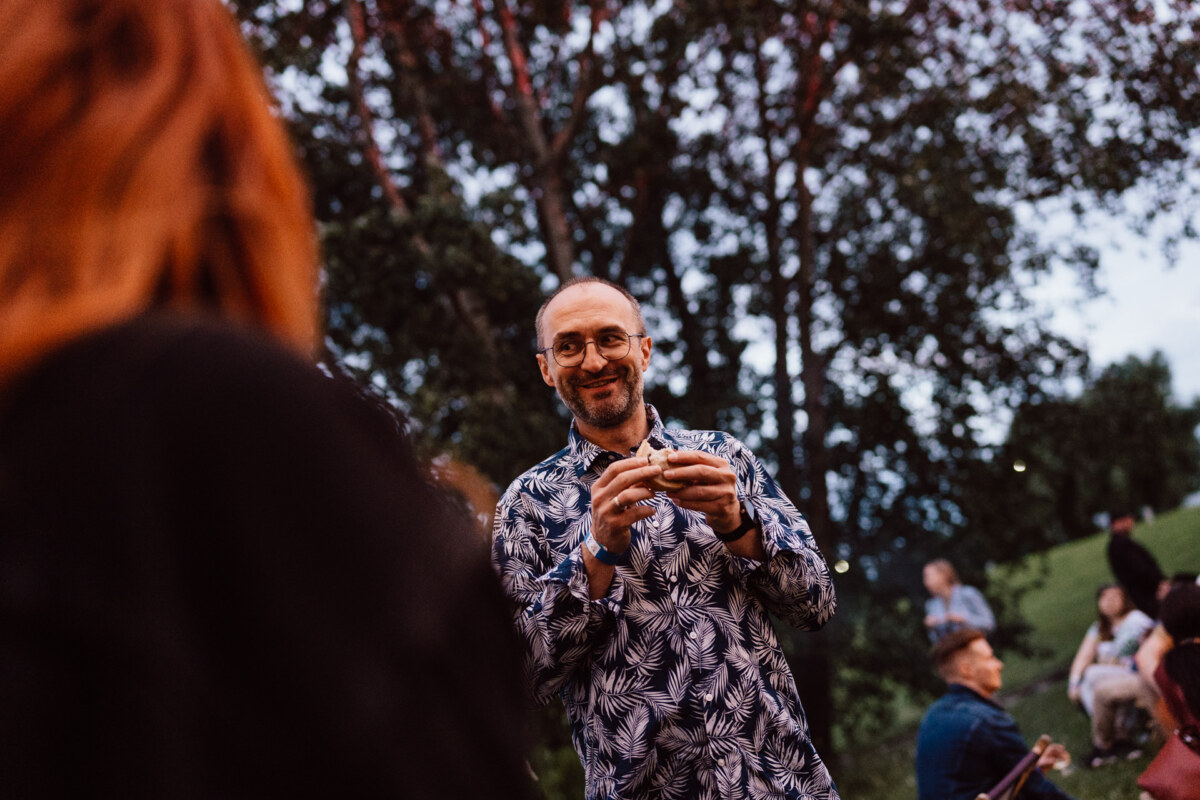 A man wearing glasses and a patterned shirt smiles and holds something in his hands at an outdoor meeting, with people and trees in the background.
